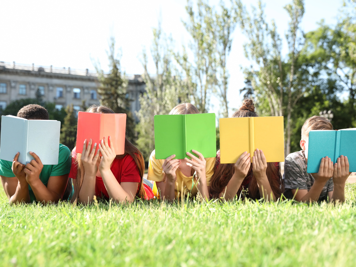 Kids laying on stomachs outside with different colored books in front of their faces.