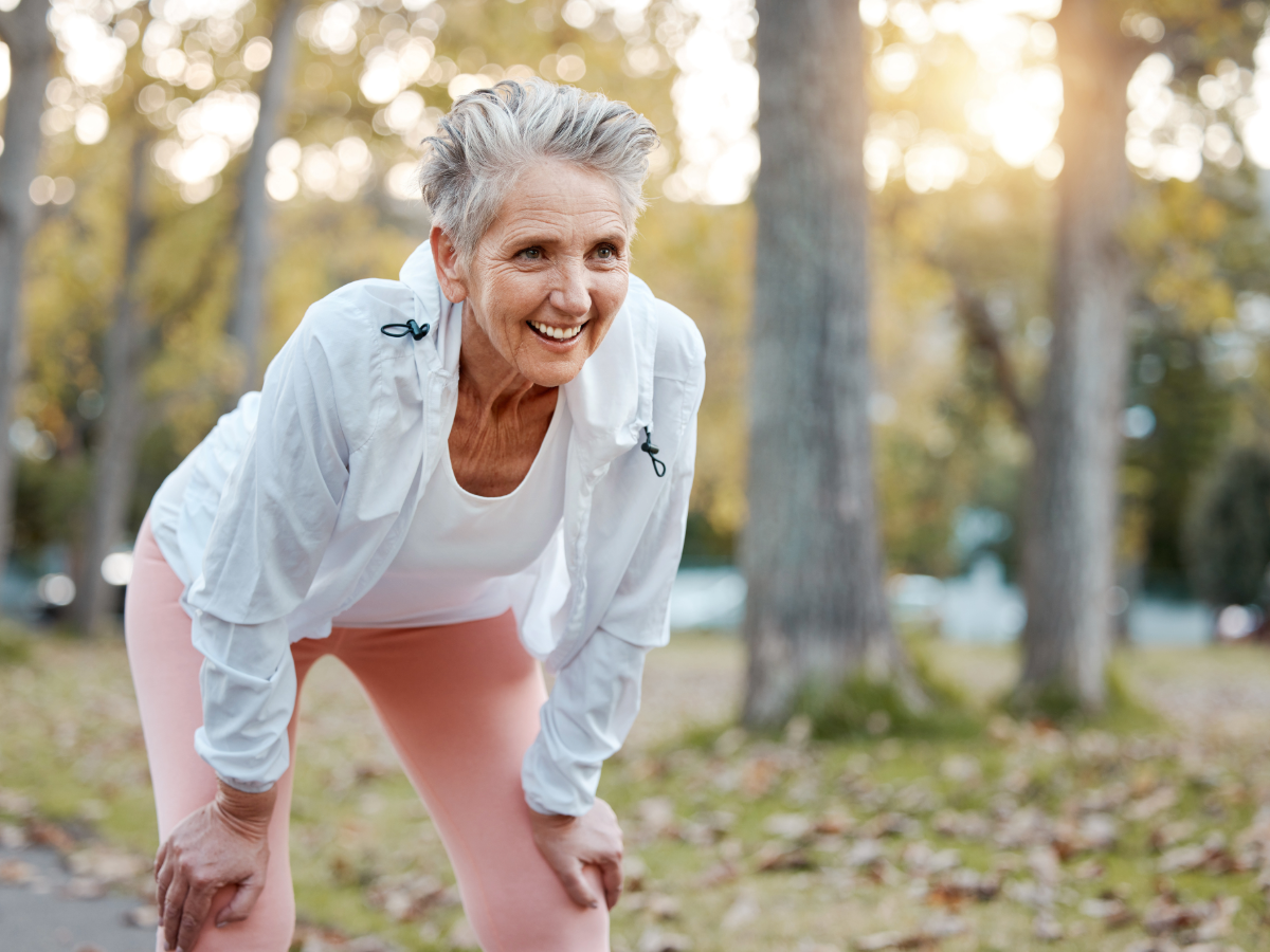 Active senior woman outdoors on jogging trail with hands on knees smiling.