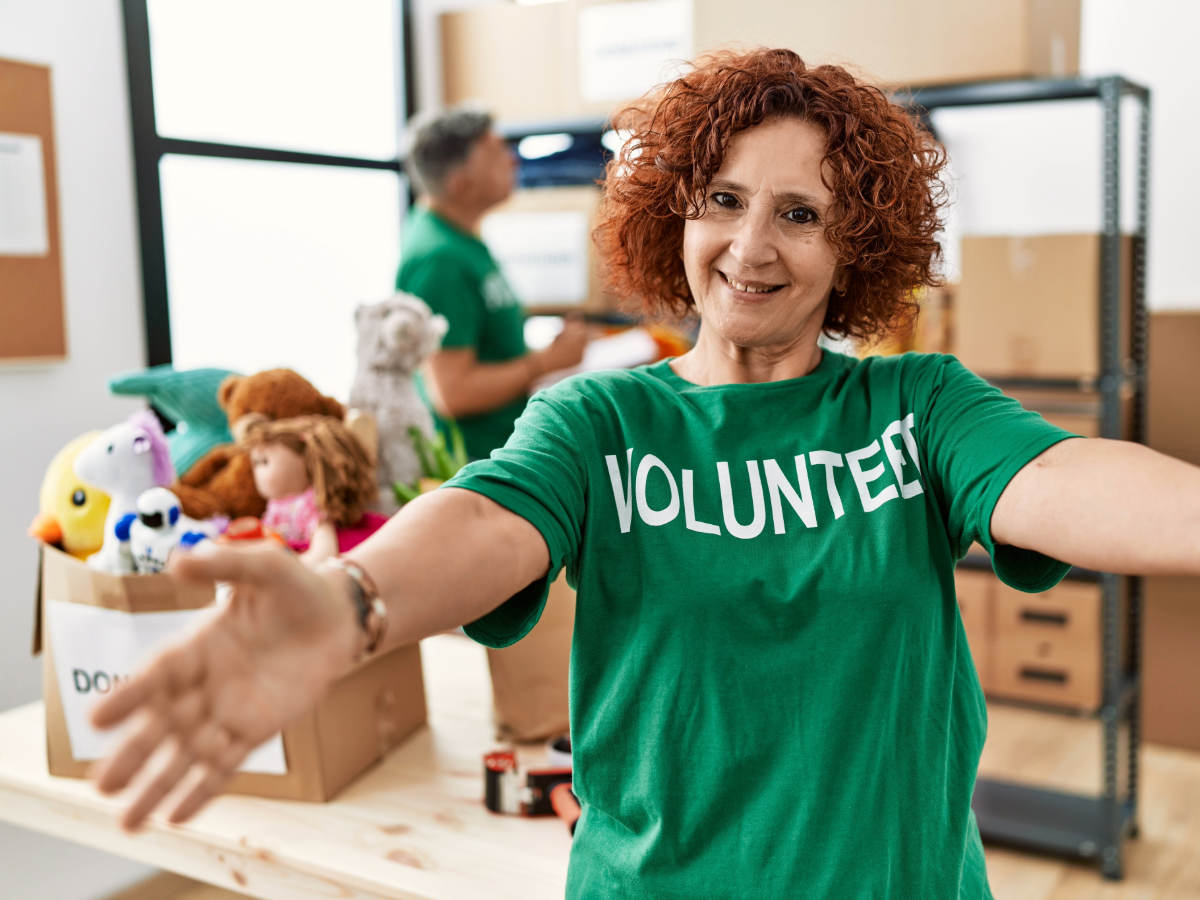 Senior woman in a green shirt that says volunteer on it standing in front of counter with boxes of donations.