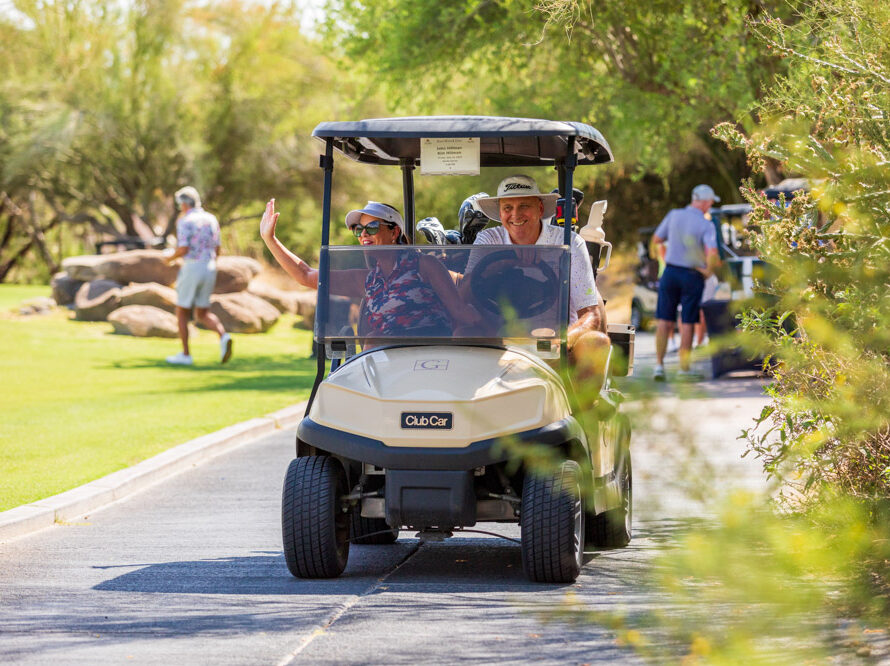 People smiling and waving while riding in a golf cart on a golf course path.