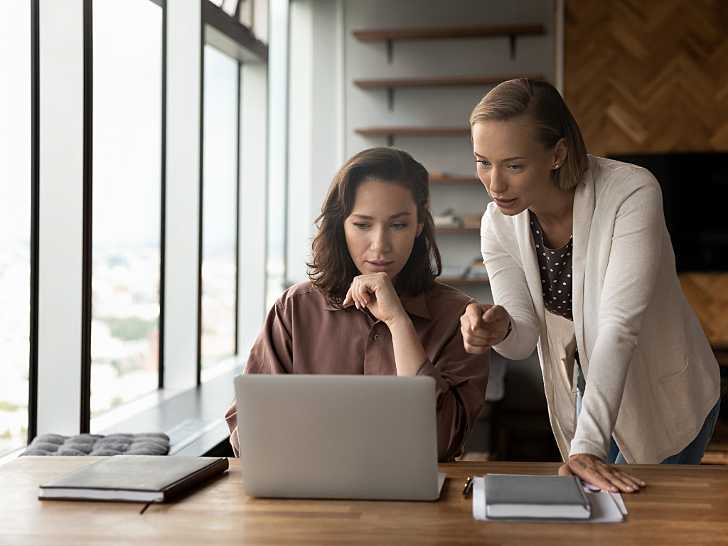 1 woman sitting at a desk with laptop while another woman is leaning over the desk pointing at the computer and mentoring the first woman.