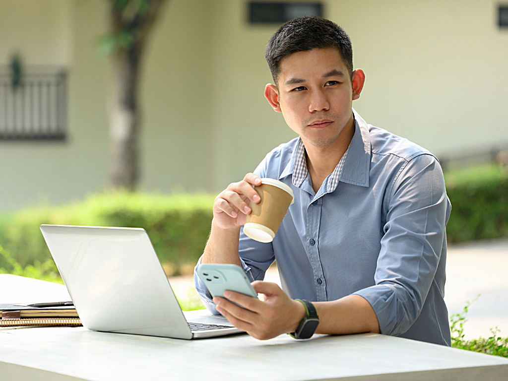 Young man sitting outdoors working remotely with coffee cup in one hand, cell phone in the other and laptop on cafe table.