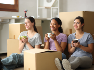 3 female roommates sitting on floor drinking coffee with moving boxes around them.