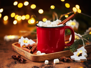 Red coffee mug filled with coffee, marshmellows and cinnamon sticks on a tray with marshmellows and cinnamon sticks around the mug and lights and pine in the background for article The 12 Best Coffee Shops in Tucson for Newcomers moving to Tucson.