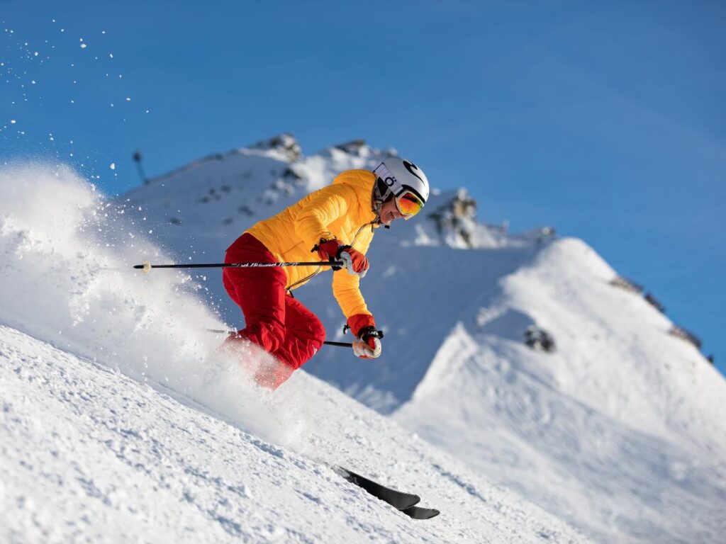 Downhill snow skier in yellow jacket and red snow pants skiing down the mountain for article Places to Ski Near Tucson for newcomers moving to Tucson.