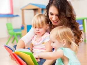 Woman with 2 young girls in her arms sitting on her lap on the floor as she holds a book out in front of all of them and 1 child is pointing at the pages for article Childcare Options in Tucson for newcomers moving to Tucson.