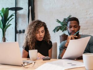 Couple sitting at kitchen table that has budget papers, coffee cups and laptop on it - The couple is looking at one of the papers and crunching numbers on a calculator for article Cost of Living in Tucson Compared to Other Major U.S. Cities